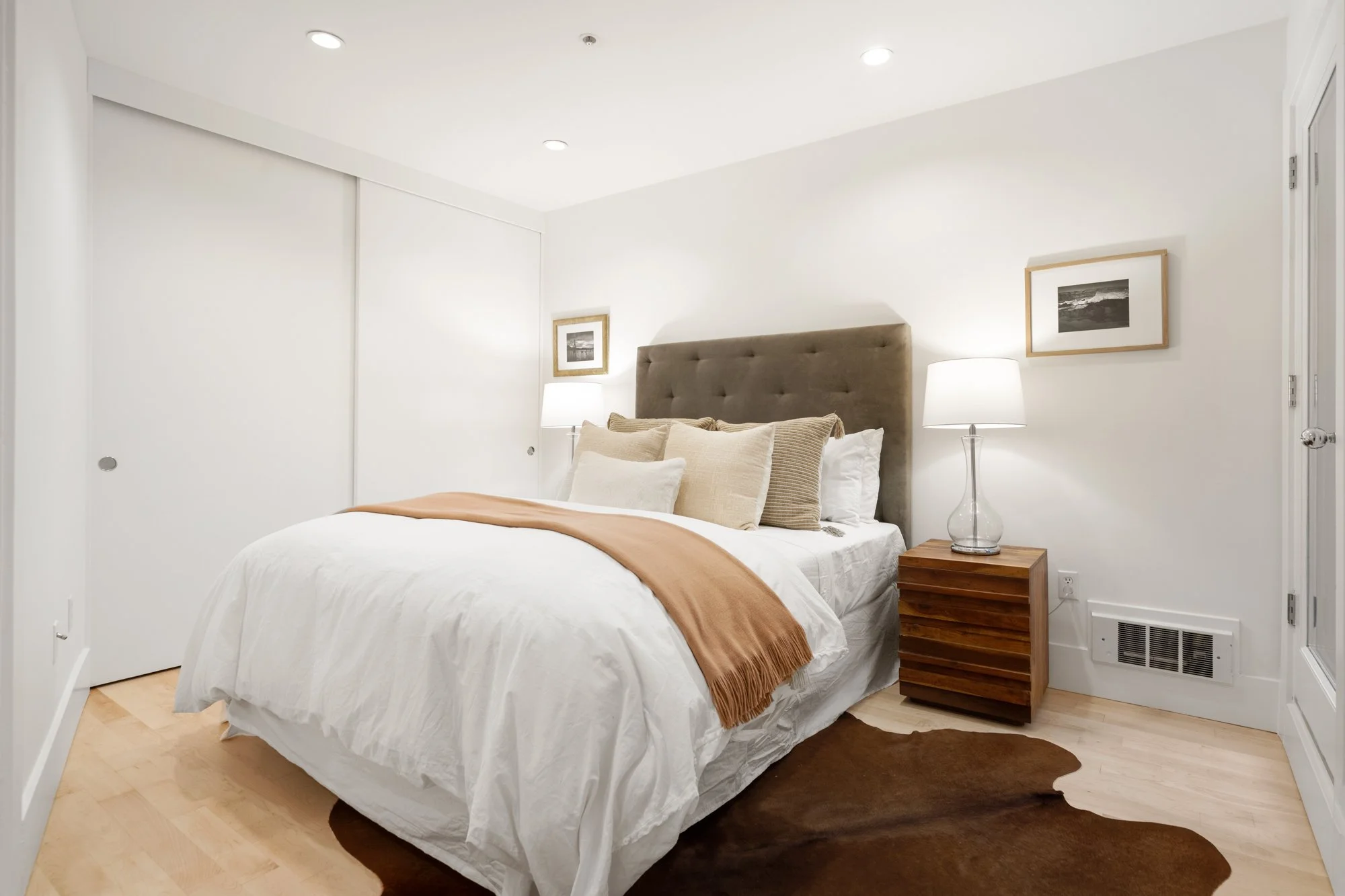 A bedroom with a bed, beige headboard, white bedding, multiple throw pillows, brown blanket, wooden nightstand, table lamp, framed black-and-white photos, brown cowhide rug, white closet doors, and a vent on the wall.