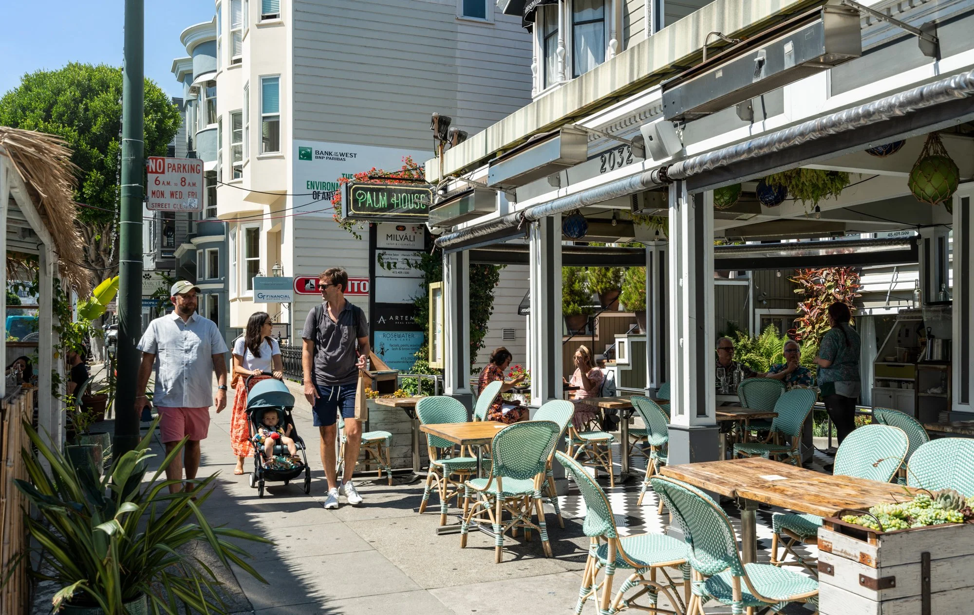 Outdoor restaurant patio with tables and chairs, people dining and walking, and a neon sign reading 'Palm House'.