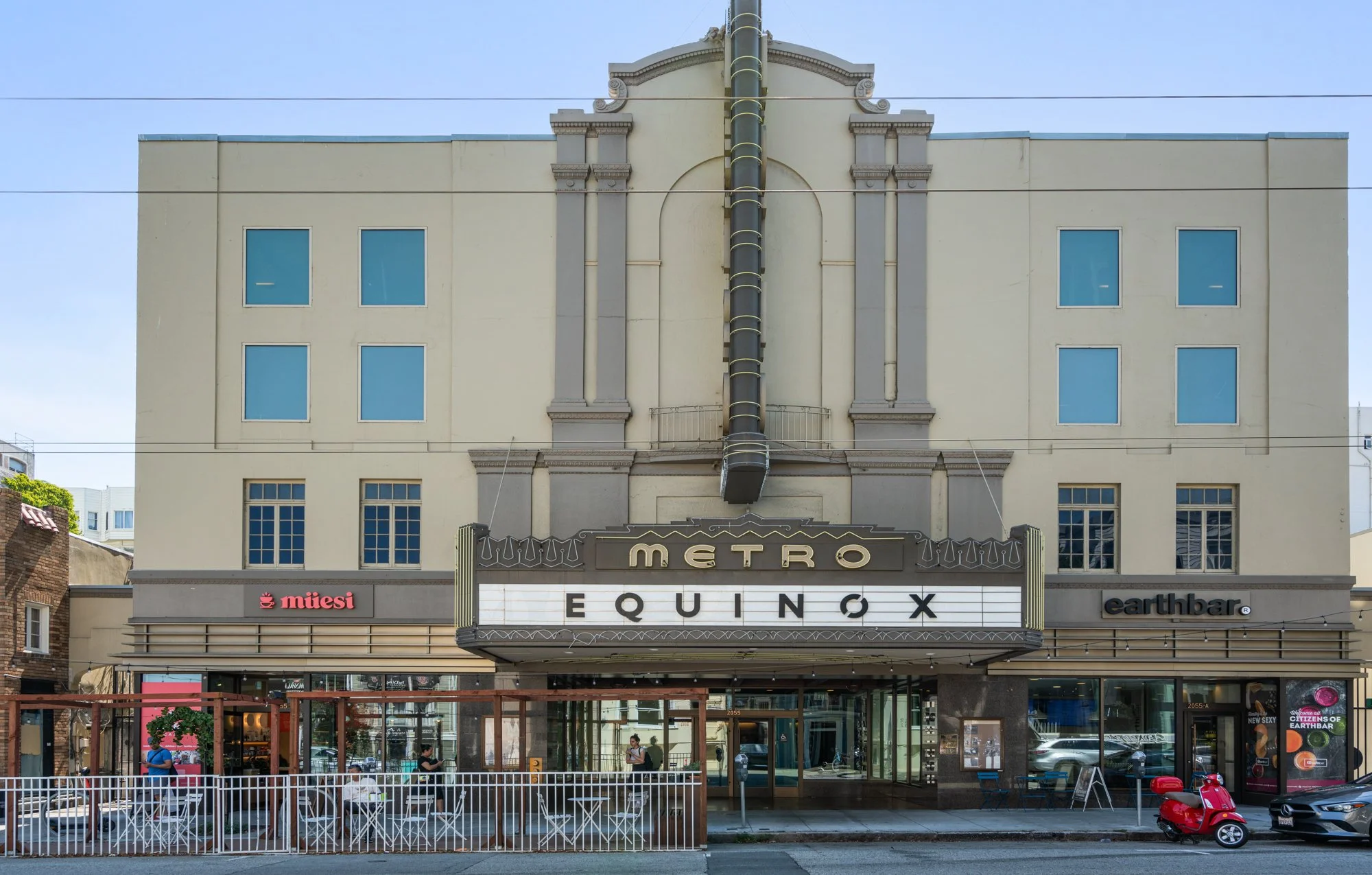 The facade of a vintage-style movie theater with a marquee reading 'METRO EQUINOX'. The theater has an art deco design with decorative columns and a black slide descending from the roof. There are storefronts for 'müesi' and 'earthbar', and people si