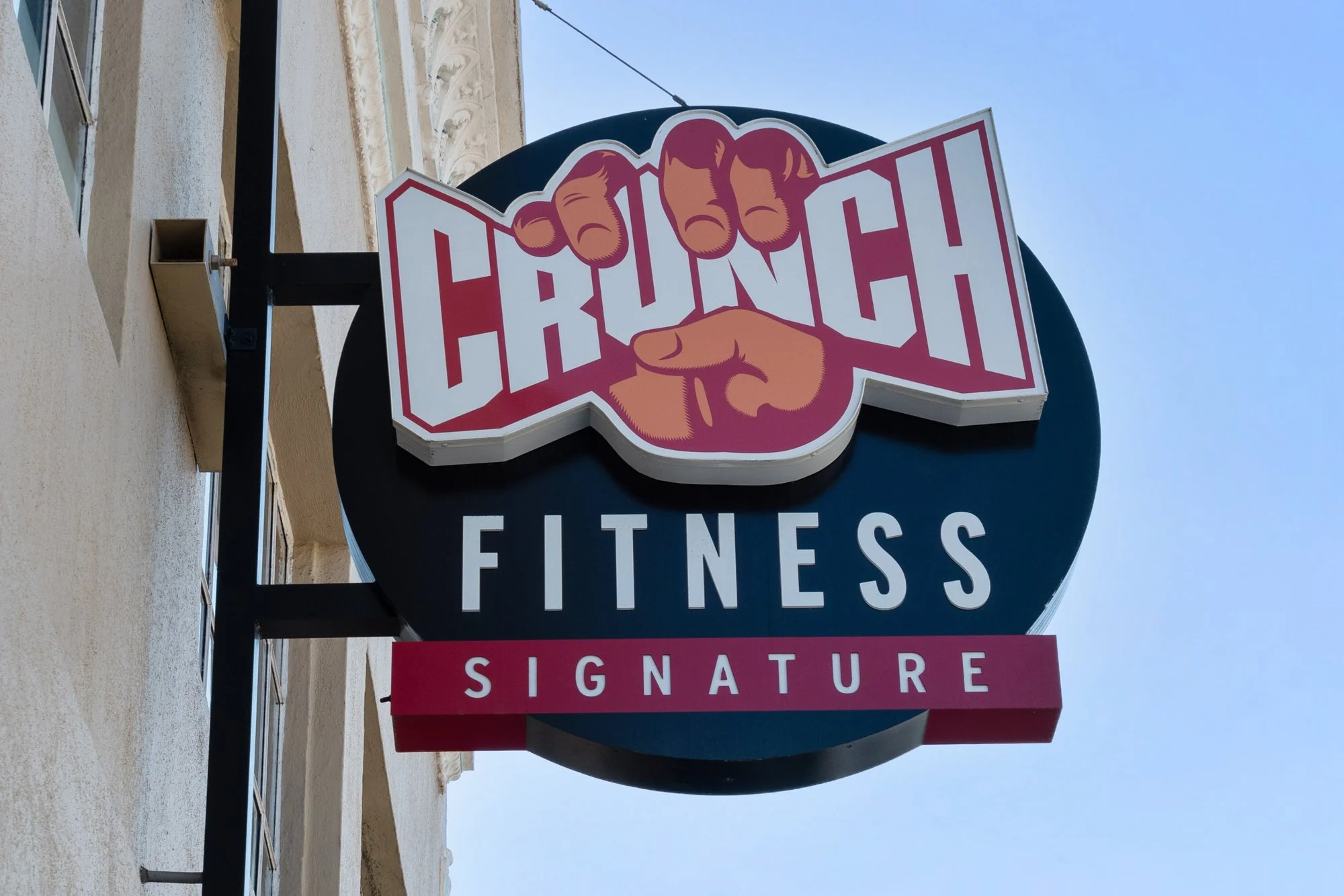 Sign for Crunch Fitness with a graphic of two fists, one pointing directly at the viewer. The sign is mounted outside a building with a clear blue sky in the background.