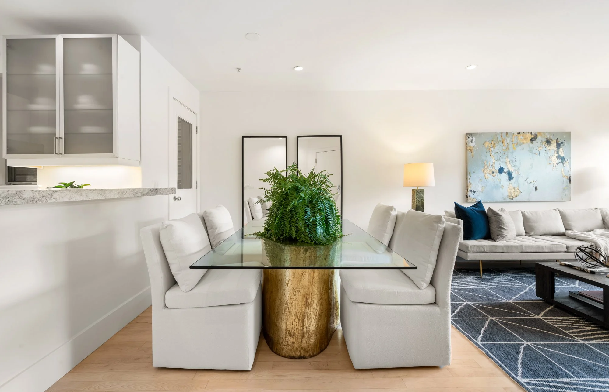 Modern living and dining room with white furniture, glass dining table, green plant centerpiece, black framed mirrors, abstract wall art, rug, and side table with lamp.