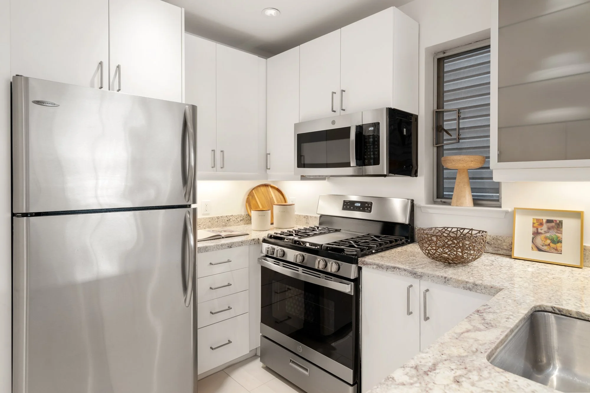 Modern kitchen with stainless steel refrigerator, stove, and microwave, white cabinets, granite countertops, and a small window with a decorative object and framed photo on the counter.
