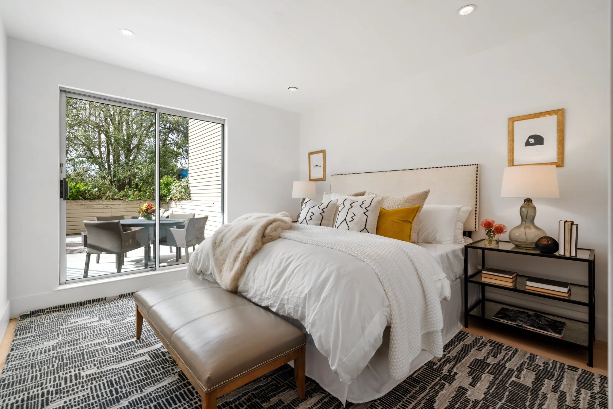 Modern bedroom with white bedding, pillows, and throw blanket, black and white patterned rug, beige bench at foot of bed, black side table with lamp, art prints on wall, sliding glass door leading to outdoor patio with table and chairs, greenery outs