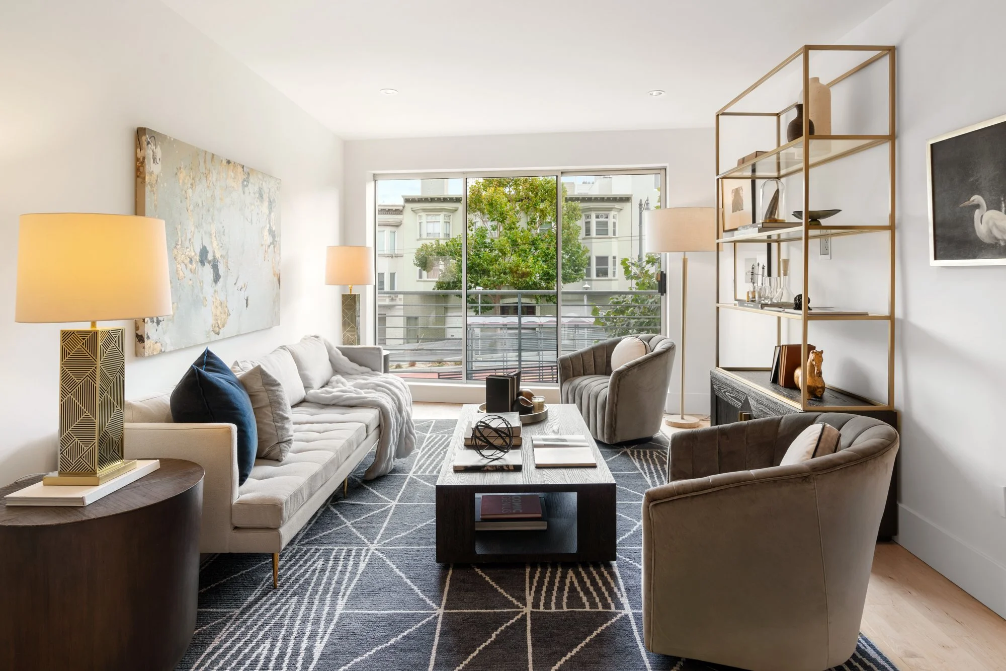 Living room with white walls, large window, sofa with pillows, armchairs, coffee table, bookshelf with decorative items, floor lamps, abstract art on wall, patterned rug.