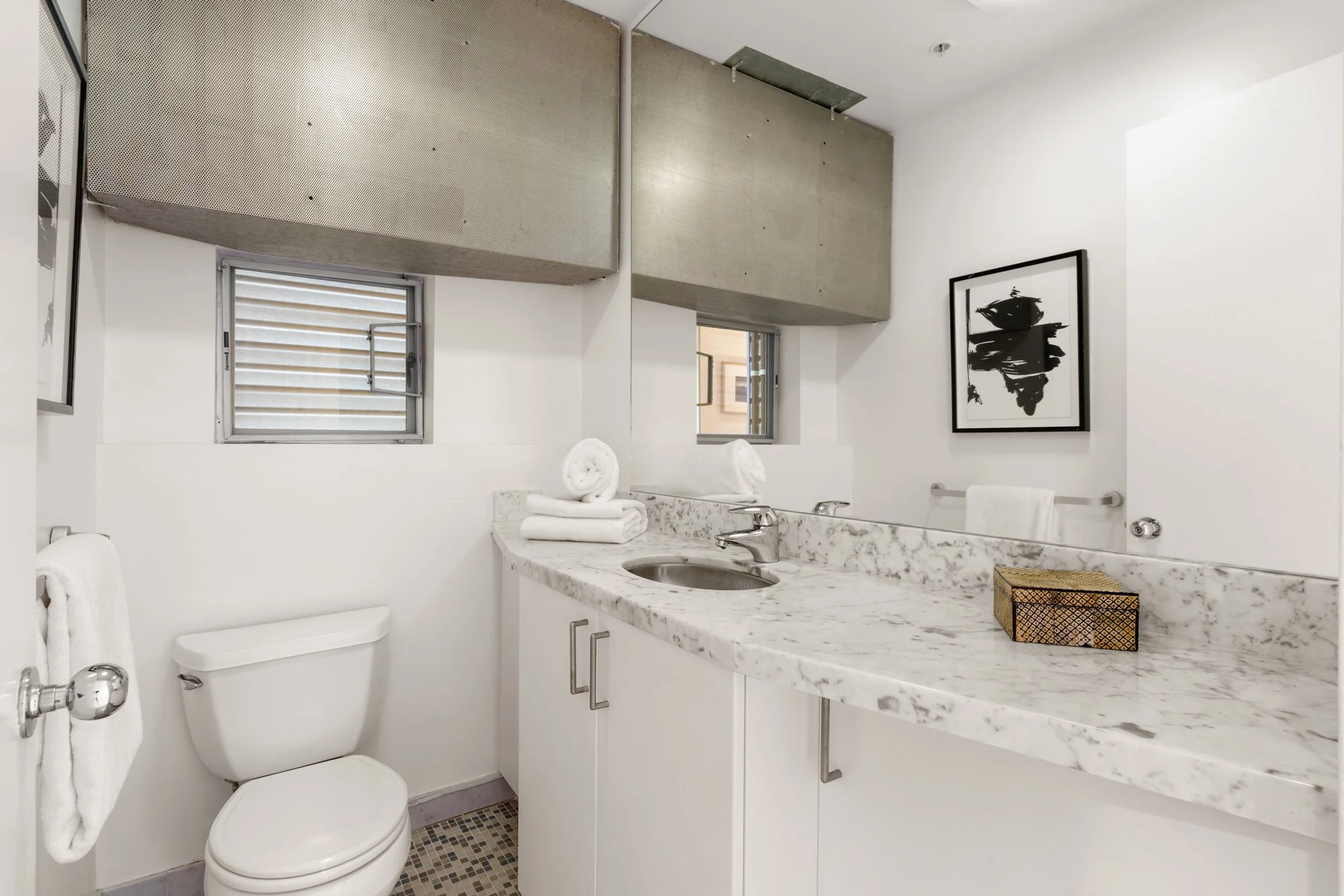 A small bathroom with a porcelain toilet, a marble countertop with a sink, and a mirror above the counter. There are folded white towels on the counter and a towel bar with a white towel next to the toilet. The bathroom has two small windows, one wit