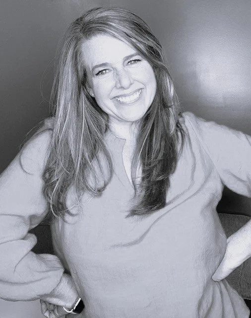 A woman smiling with her hands on her hips, wearing a long-sleeve shirt, in front of a plain wall.