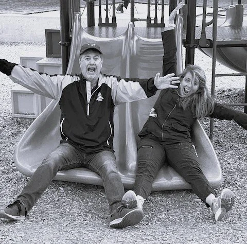 Two people, a man and a woman, sitting at the bottom of a playground slide with their arms raised and smiling.