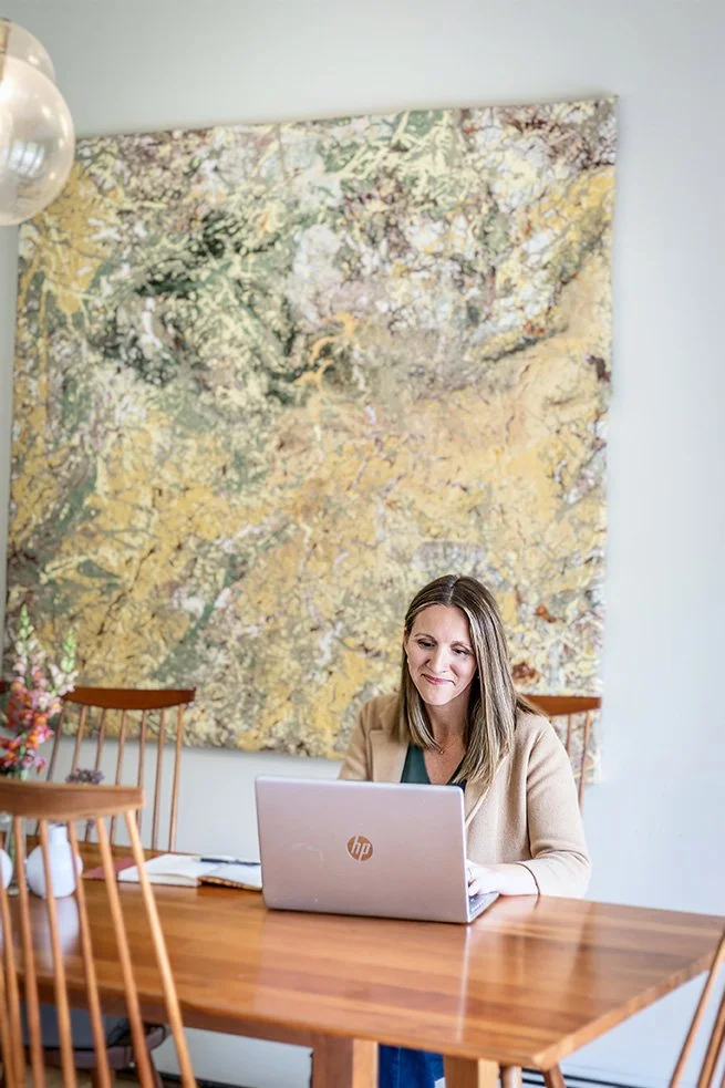 A woman sitting at a wooden table working on a laptop, with a large, colorful abstract painting on the wall behind her.