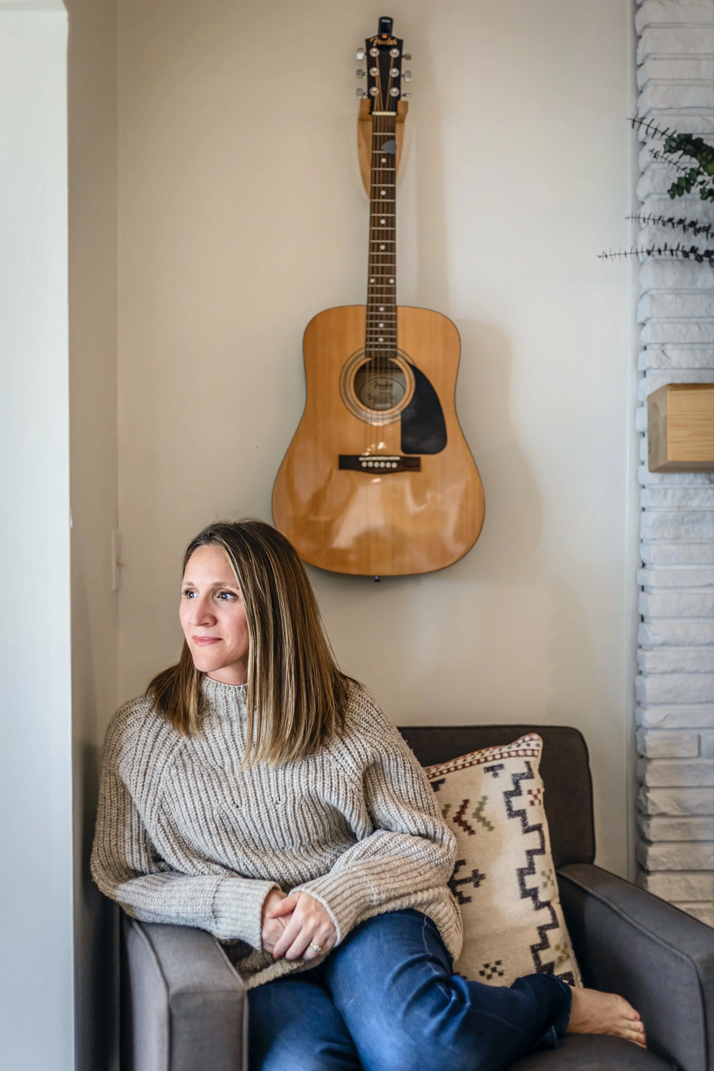 A woman sitting on a gray armchair with patterned pillow, next to a white brick wall with a large wooden acoustic guitar hanging on the wall behind her.