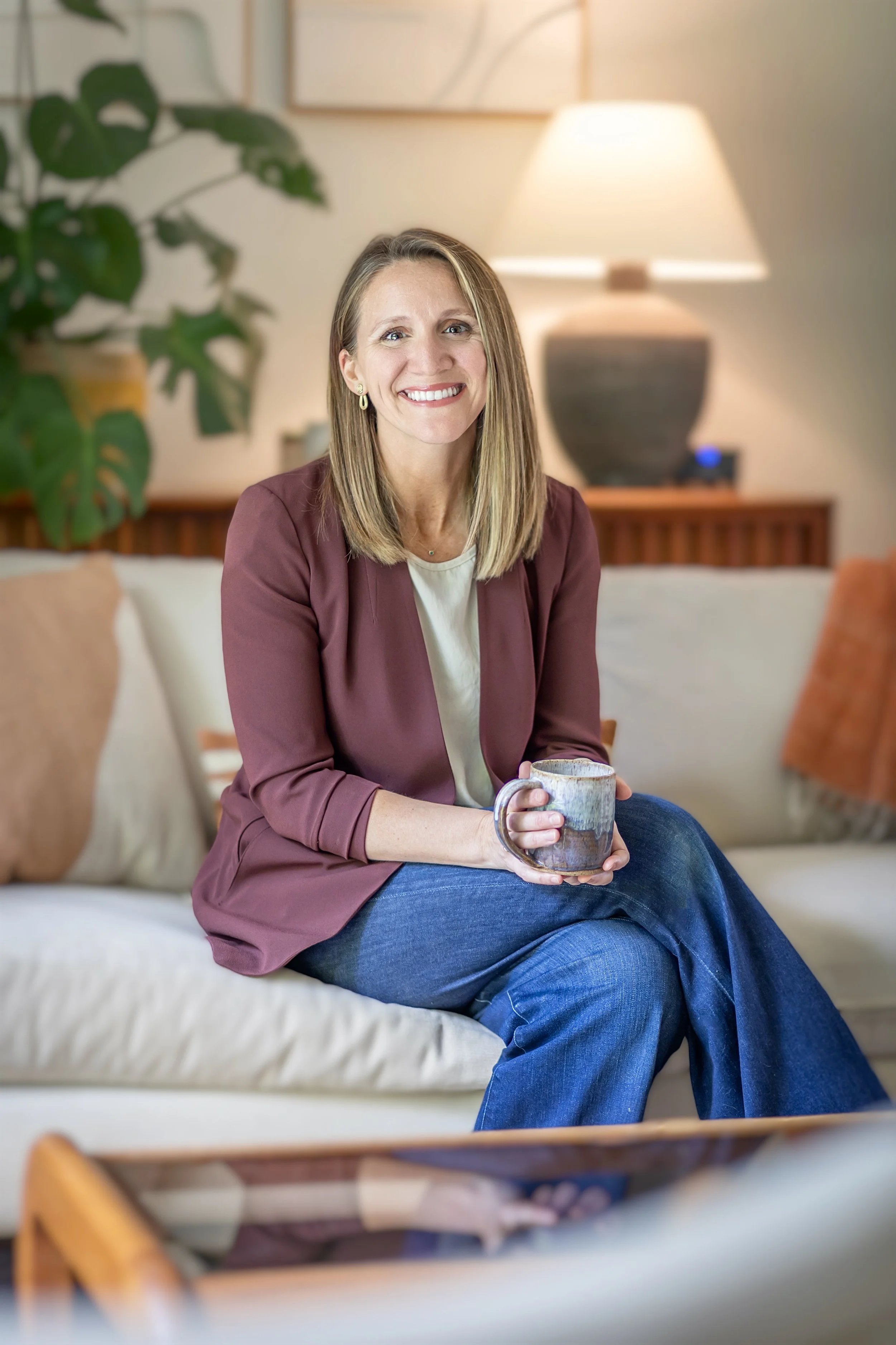 A woman with shoulder-length blonde hair, smiling, sitting on a sofa, holding a ceramic mug, wearing a maroon blazer and blue jeans, in a cozy living room with a large plant, a lamp, and framed art on the wall in the background.