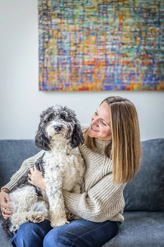 A woman with straight light brown hair, wearing a beige sweater, smiling and holding a black and white curly-haired dog on a gray couch. There is an abstract colorful painting on the wall behind them.