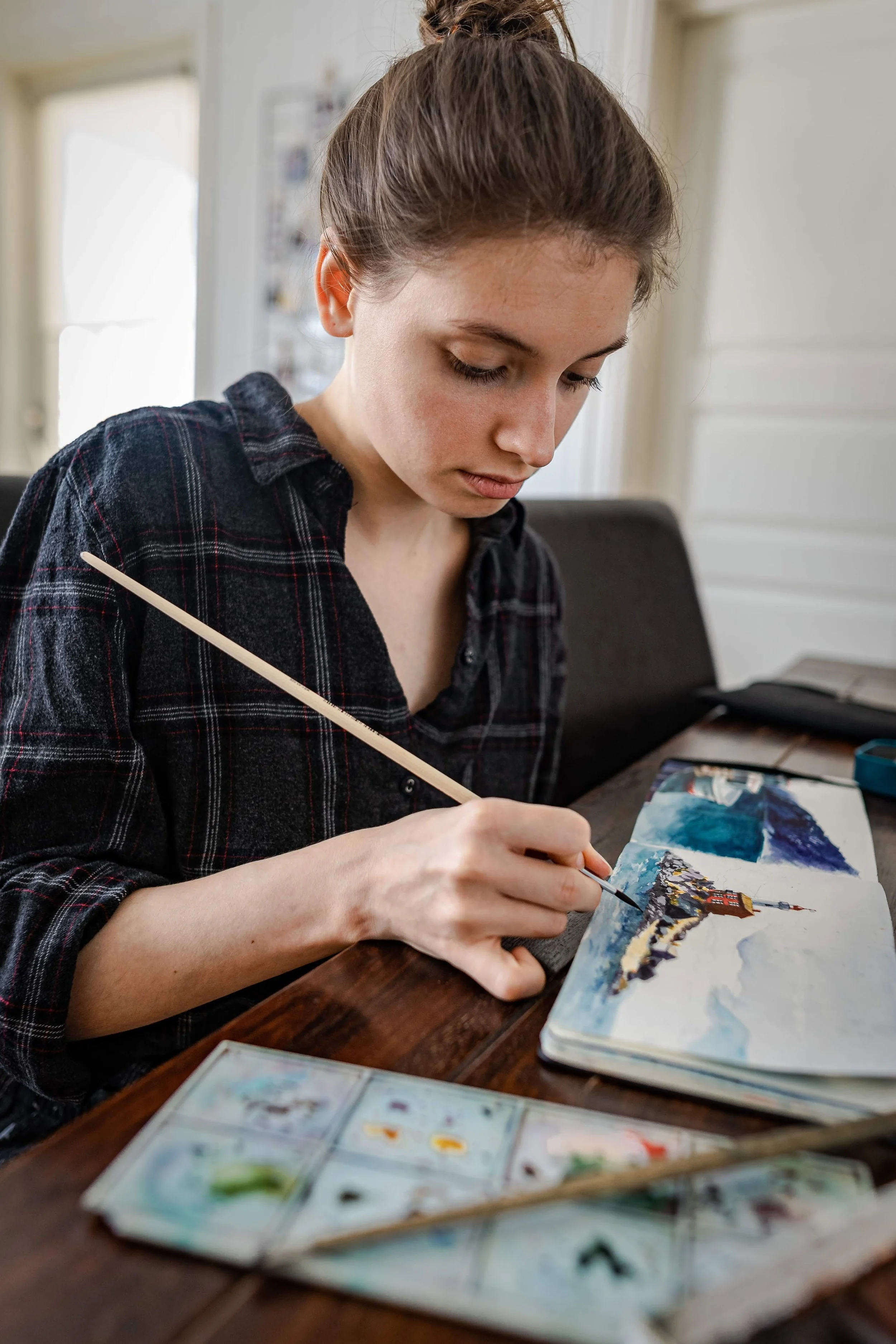 A young woman with brown hair painting a landscape scene in a sketchbook, sitting at a wooden table.