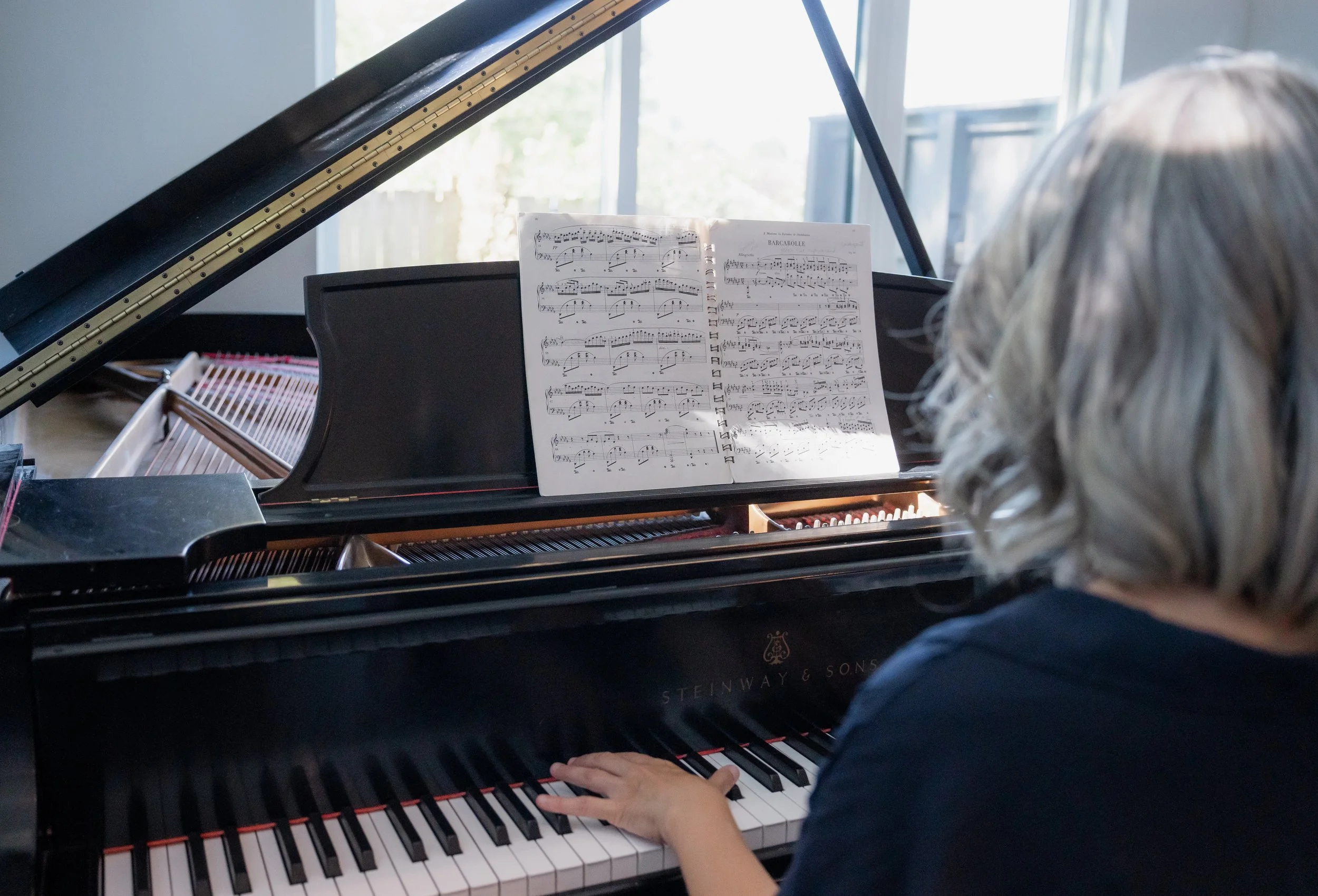 Person with gray hair playing a grand piano with sheet music on the stand, in a room with large windows.