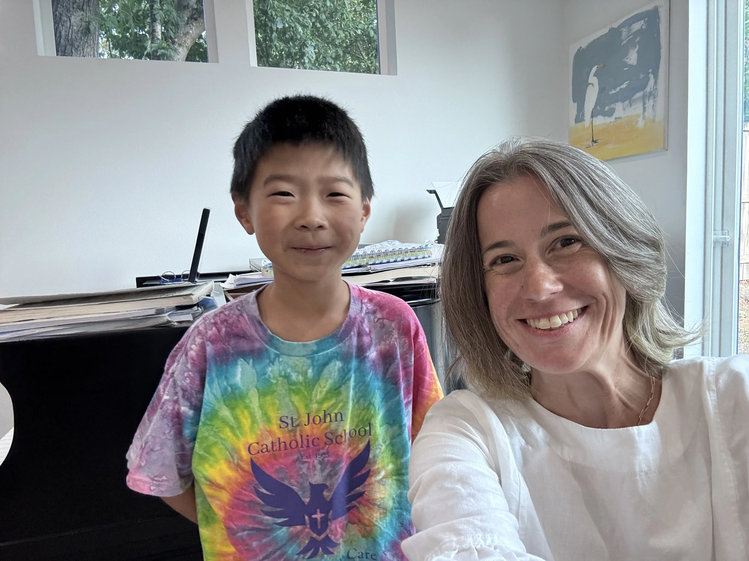 A woman and a boy smiling for a selfie in a room with white walls and large windows, with a painting of a heron in the background. The boy is wearing a colorful tie-dye shirt with the words 'St. John Catholic School' and a phoenix logo.