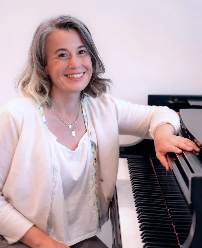 A woman, Heather Howland, with shoulder-length gray hair and a bright smile sits at a piano, wearing a light-colored cardigan and jewelry.