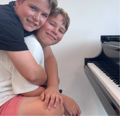 Two young boys smiling, hugging, and sitting near a piano.