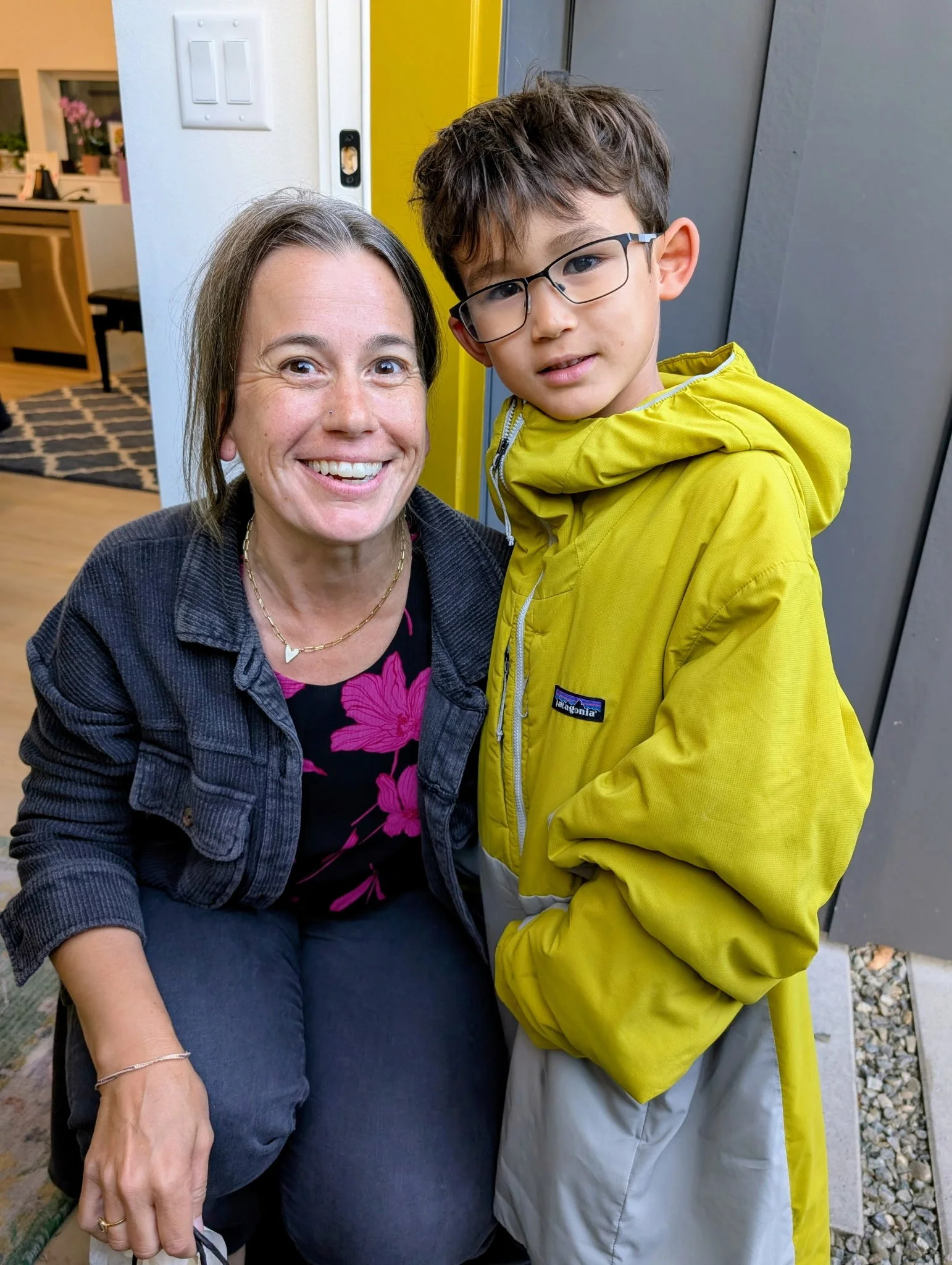 A woman with brown hair and a young boy with glasses and brown hair smiling together inside near a door