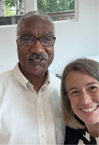 A close-up photo of two smiling people, a man with glasses and a woman, inside a room with large windows showing trees outside.