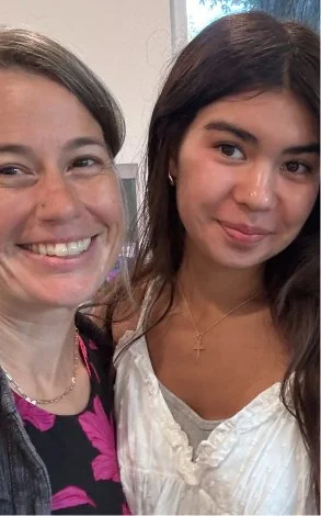 Two women taking a selfie indoors, smiling, with a window in the background.