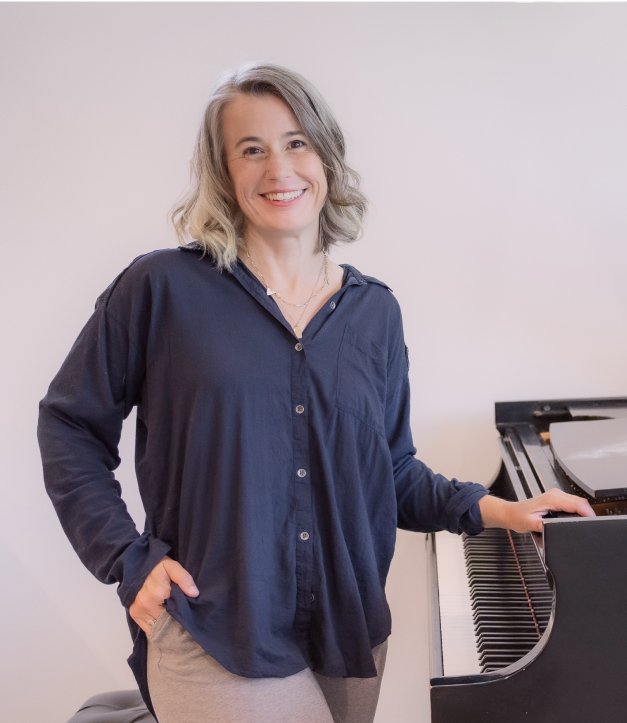 Woman, Heather Howland, in navy shirt and beige pants standing next to a grand piano in a room with white walls, smiling at the camera.