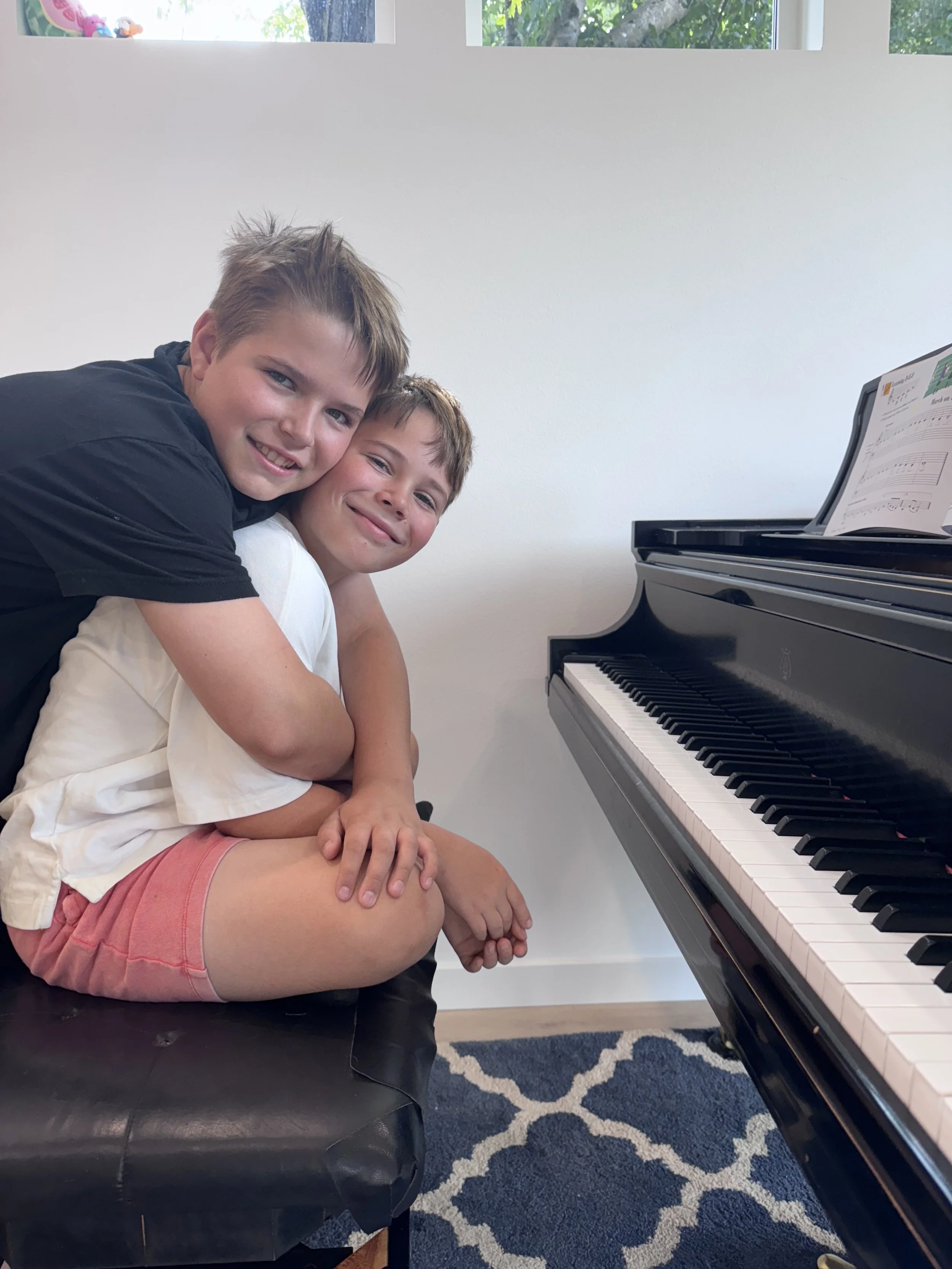 Two boys sitting on a piano bench next to a black grand piano, smiling and hugging each other, with sheet music on the piano.