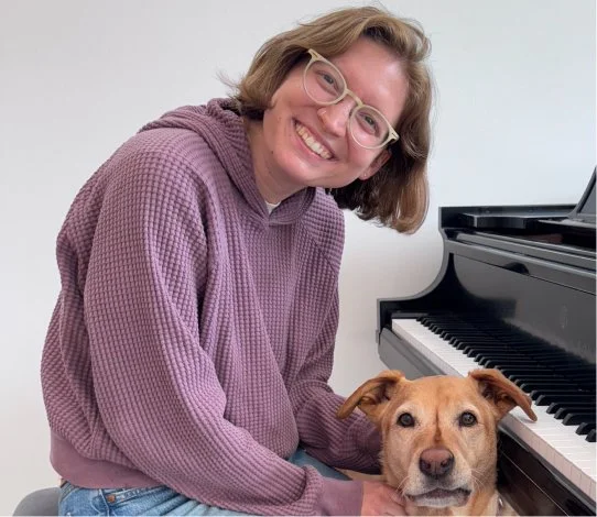A smiling woman with glasses wearing a purple hoodie sitting beside a tan dog near a piano.