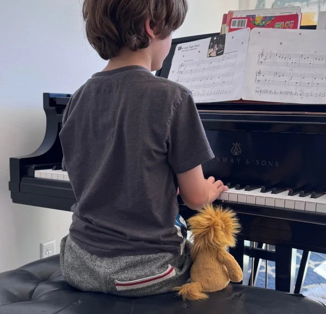 A young boy sitting at a piano, playing music, with a plush lion toy beside him and sheet music on the piano.