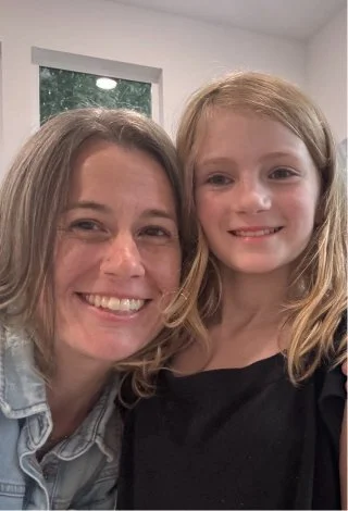 A woman and a young girl smiling and taking a selfie inside a room with a window in the background.
