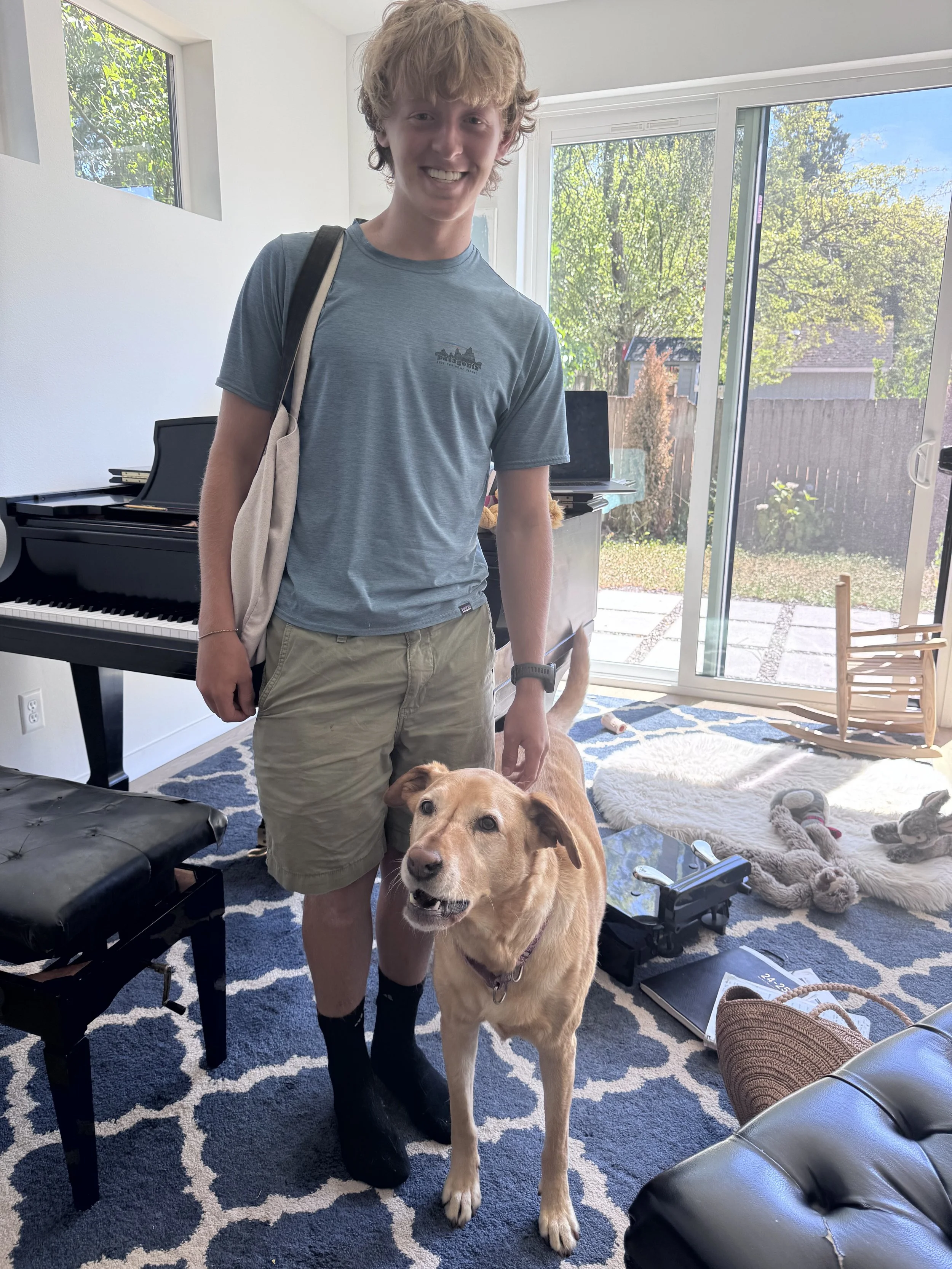 A young man with curly blonde hair, wearing a blue T-shirt, beige shorts, and black socks, standing inside a living room with a light-colored dog. The room has large windows and a sliding glass door leading to a backyard with trees. There is a black 