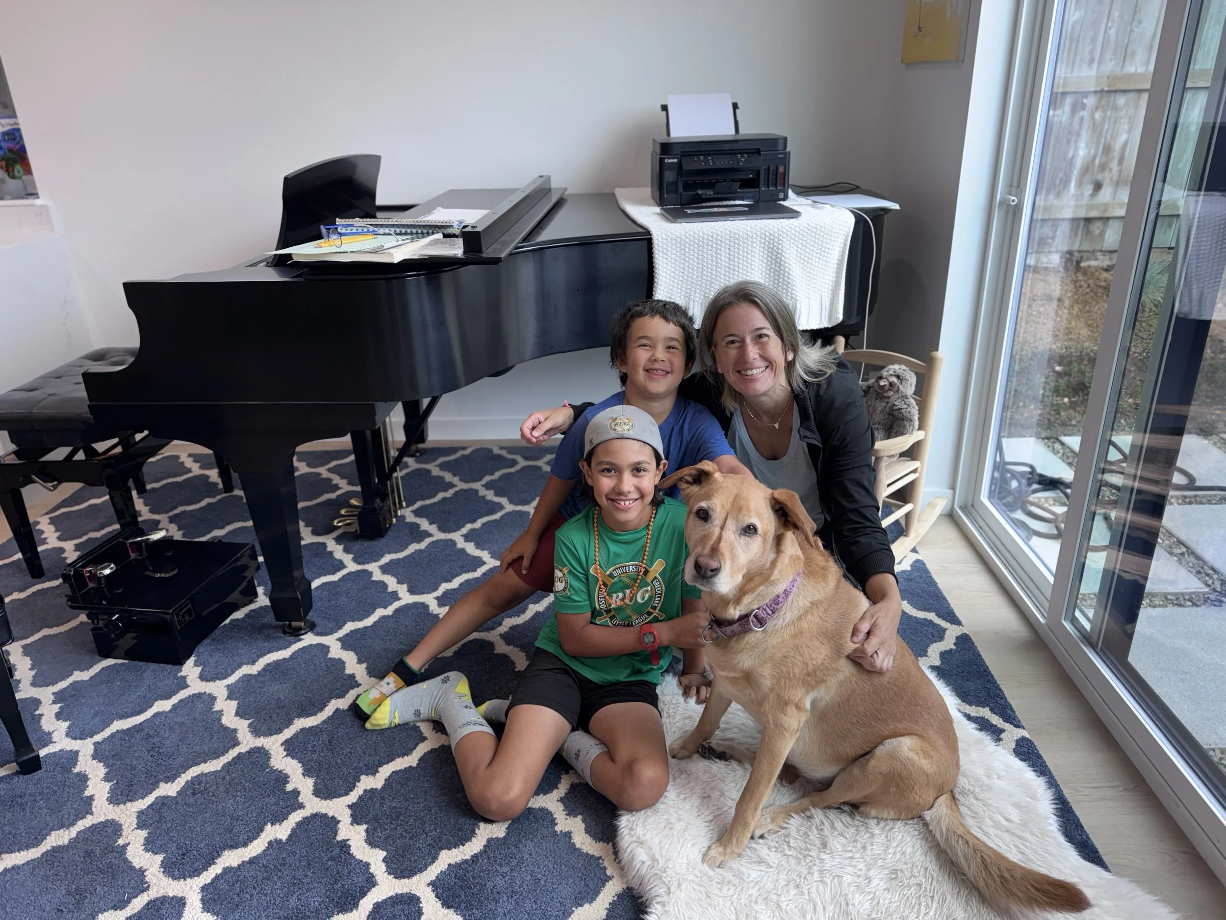 A woman and two young boys sitting on a white fluffy rug indoors, smiling with a large tan dog. Piano and printer in the background, near a sliding glass door showing a backyard.