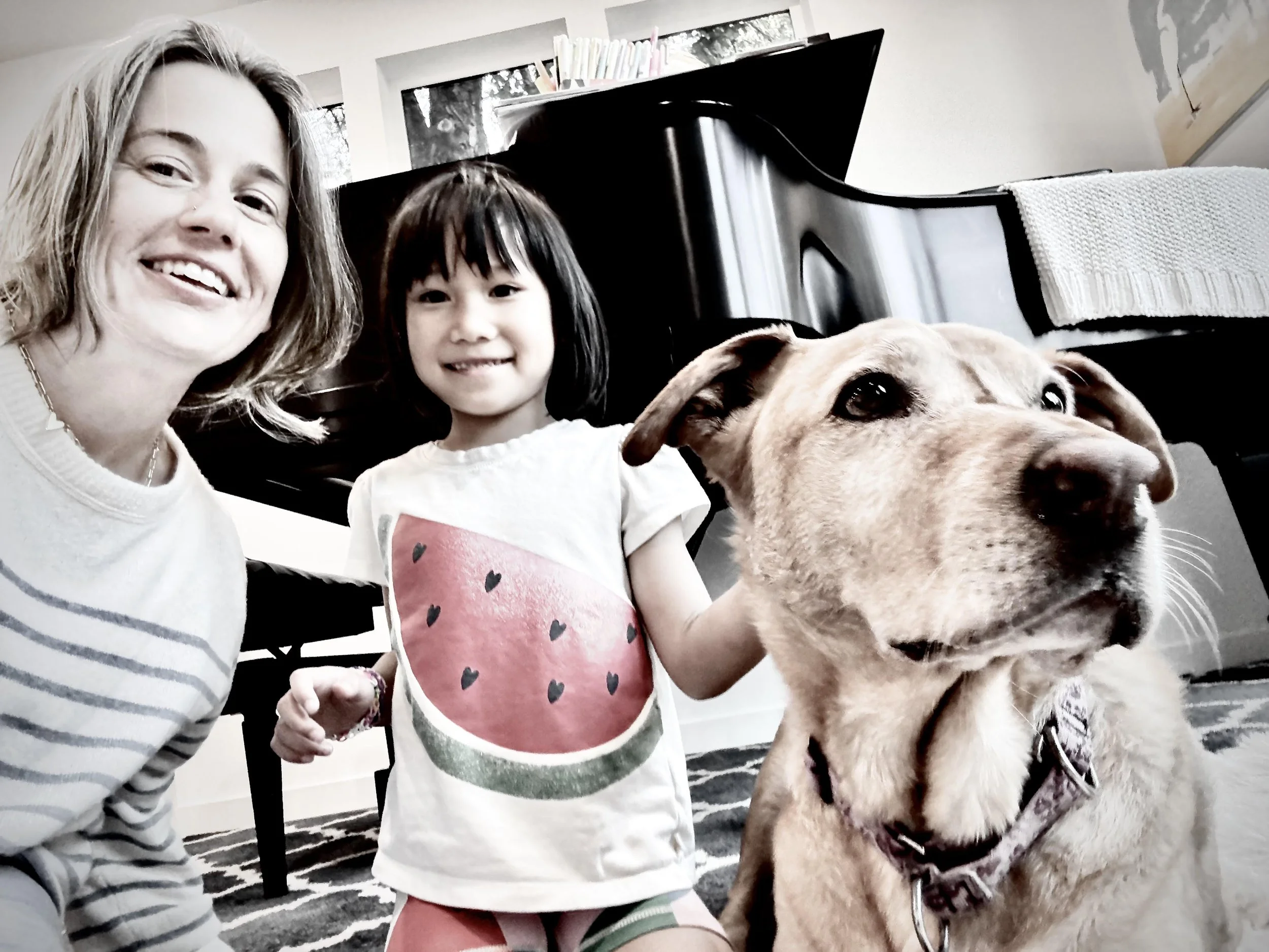 A woman, a young girl, and a large dog taking a selfie together indoors near a piano.