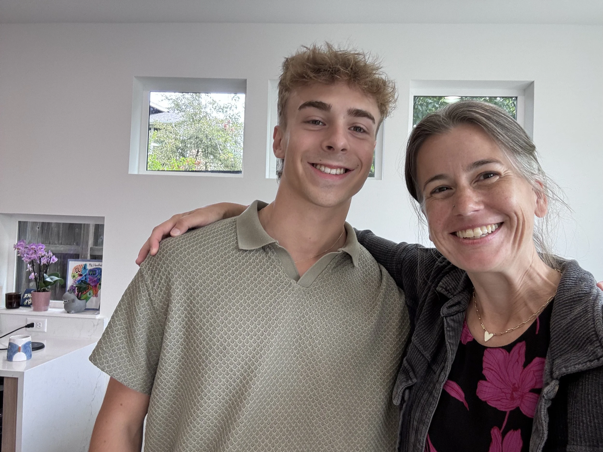 A woman and a young man smiling and posing for a selfie indoors. The woman has gray hair tied back and is wearing a necklace with a heart pendant and a floral top. The young man has curly blond hair and is wearing a beige textured polo shirt. They ar