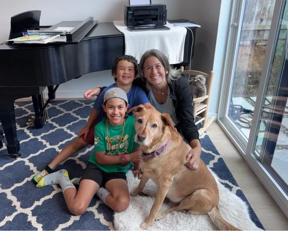 A woman and two children sitting on a patterned rug indoors with a dog, a piano, and a sliding glass door in the background.