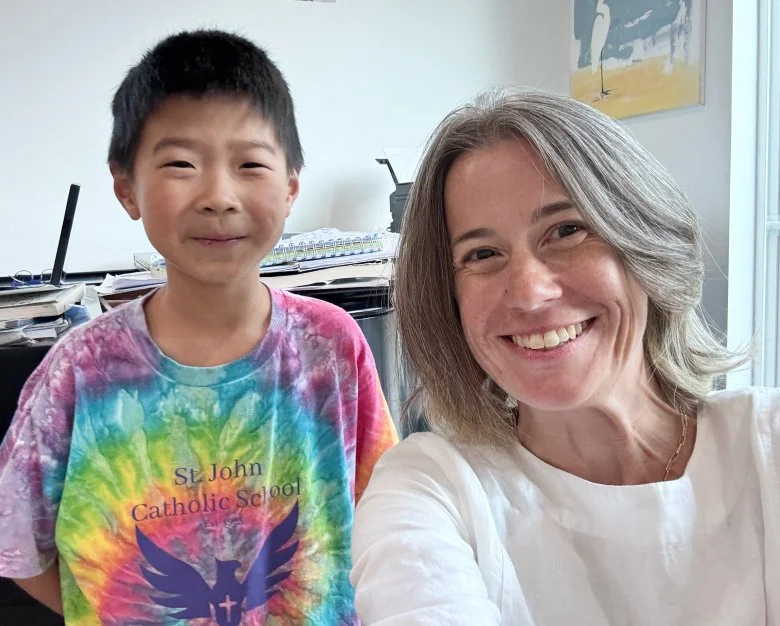 A smiling woman with shoulder-length gray hair and a boy wearing a colorful tie-dye shirt, indoors with a piano and artwork in the background.