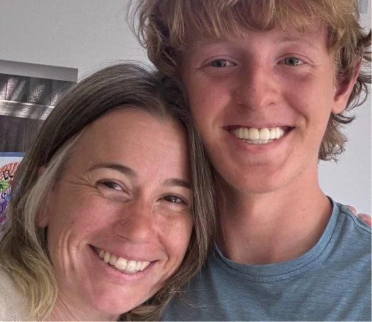 Close-up of a smiling woman with long hair and a young man with curly hair, both showing teeth, indoors.