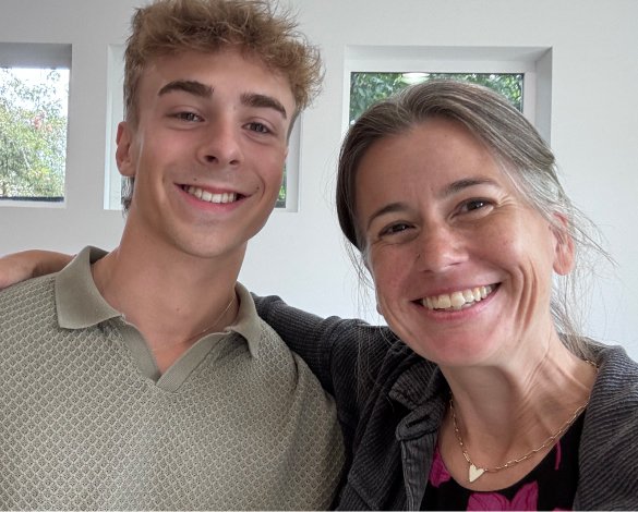 A young man and an older woman smiling and taking a selfie together inside a house with windows in the background.