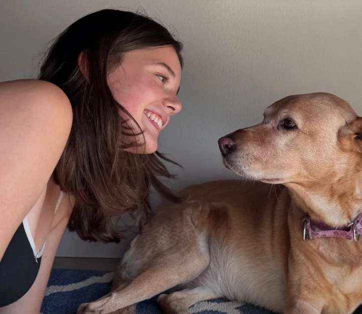 A young woman and a dog face each other indoors, lying on the floor.
The woman has shoulder-length dark hair and is smiling at the dog.
The dog is a tan-colored breed, wearing a purple collar, and looking at the woman.