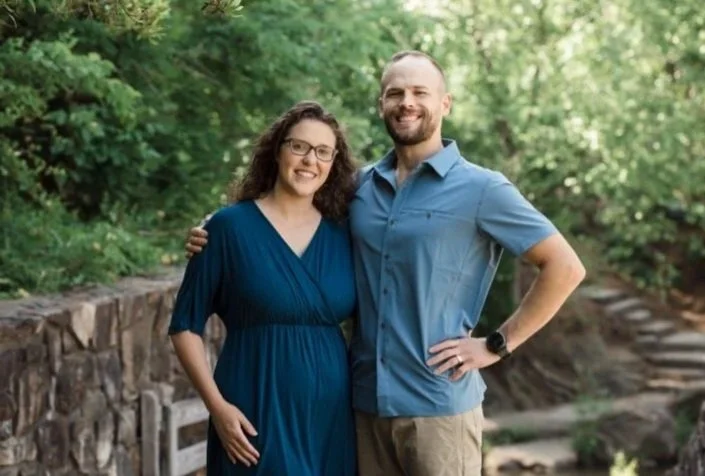 A smiling couple standing outdoors on a path surrounded by lush green trees, with the woman's arm around the man's waist and the man's hand on his hip.