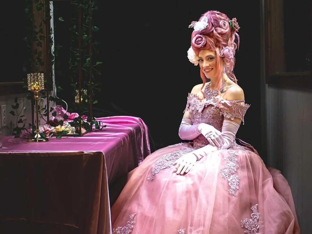 A woman dressed in an elaborate pink gown with lace and embroidery, wearing long gloves and a large, floral hairstyle, sitting at a table with pink tablecloth and decorative items, smiling in a dimly lit room.