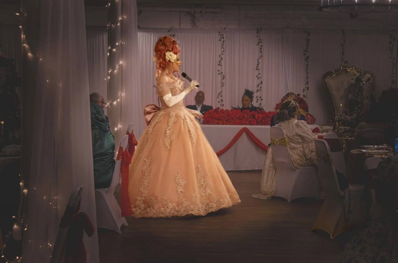 A woman in an elaborate peach gown with lace details and long gloves is speaking into a microphone at a decorated event, with seated attendees in formal attire and a backdrop of white curtains and string lights.