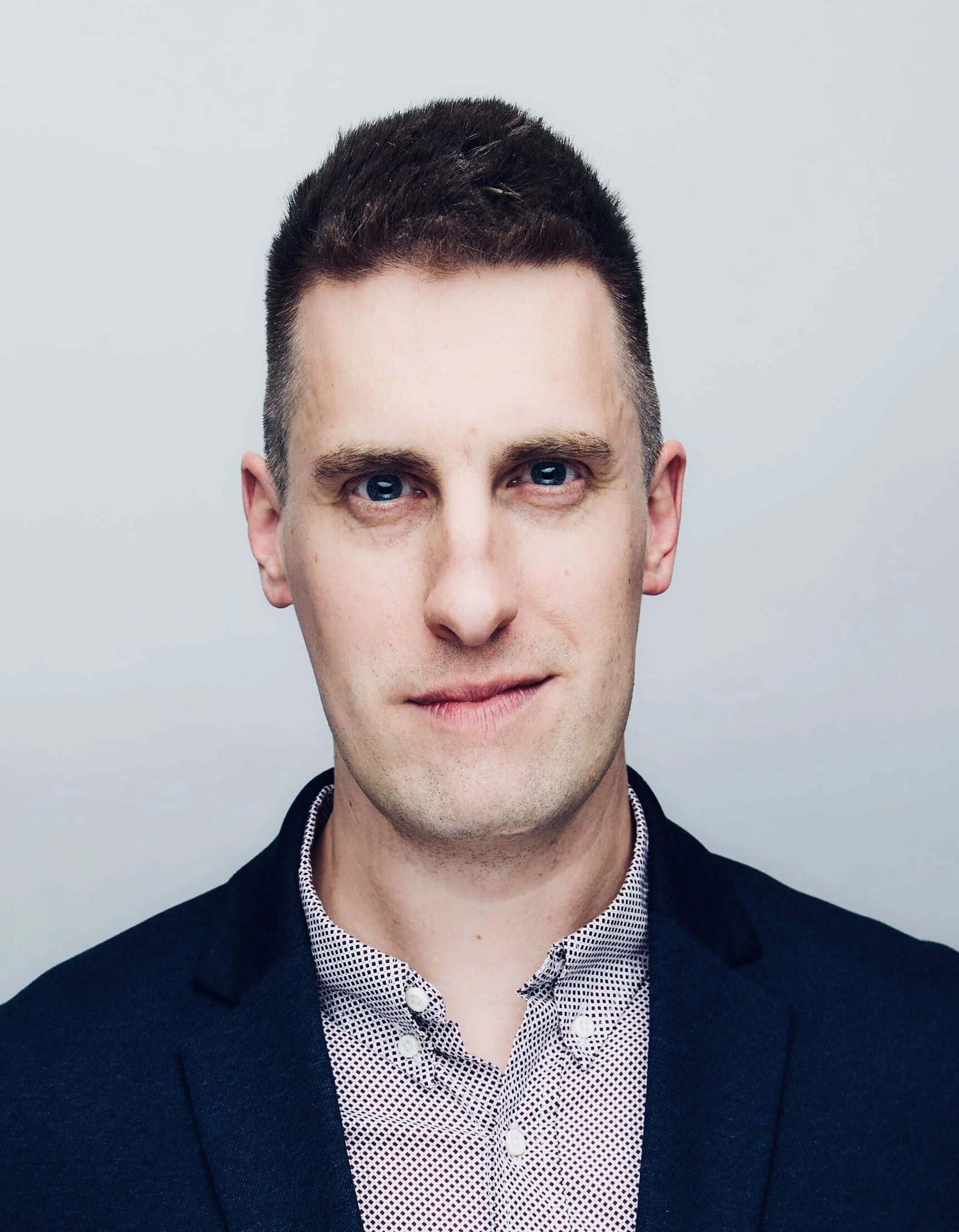 Headshot of a man with short dark hair, blue eyes, wearing a dark blazer over a patterned shirt, against a plain light background.