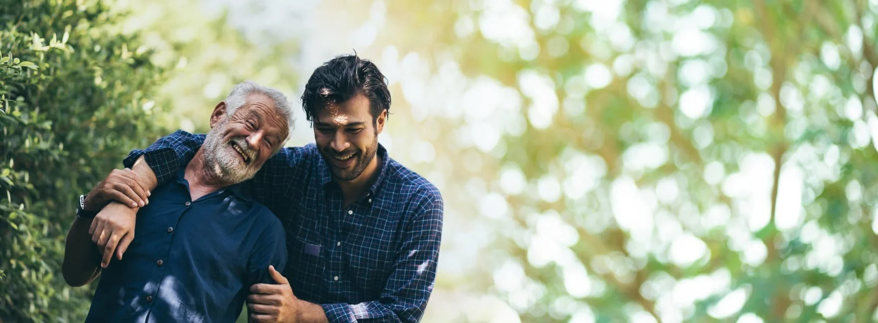 Two men, one elderly and one young, smiling and embracing outdoors in a wooded area with sunlight filtering through the trees.