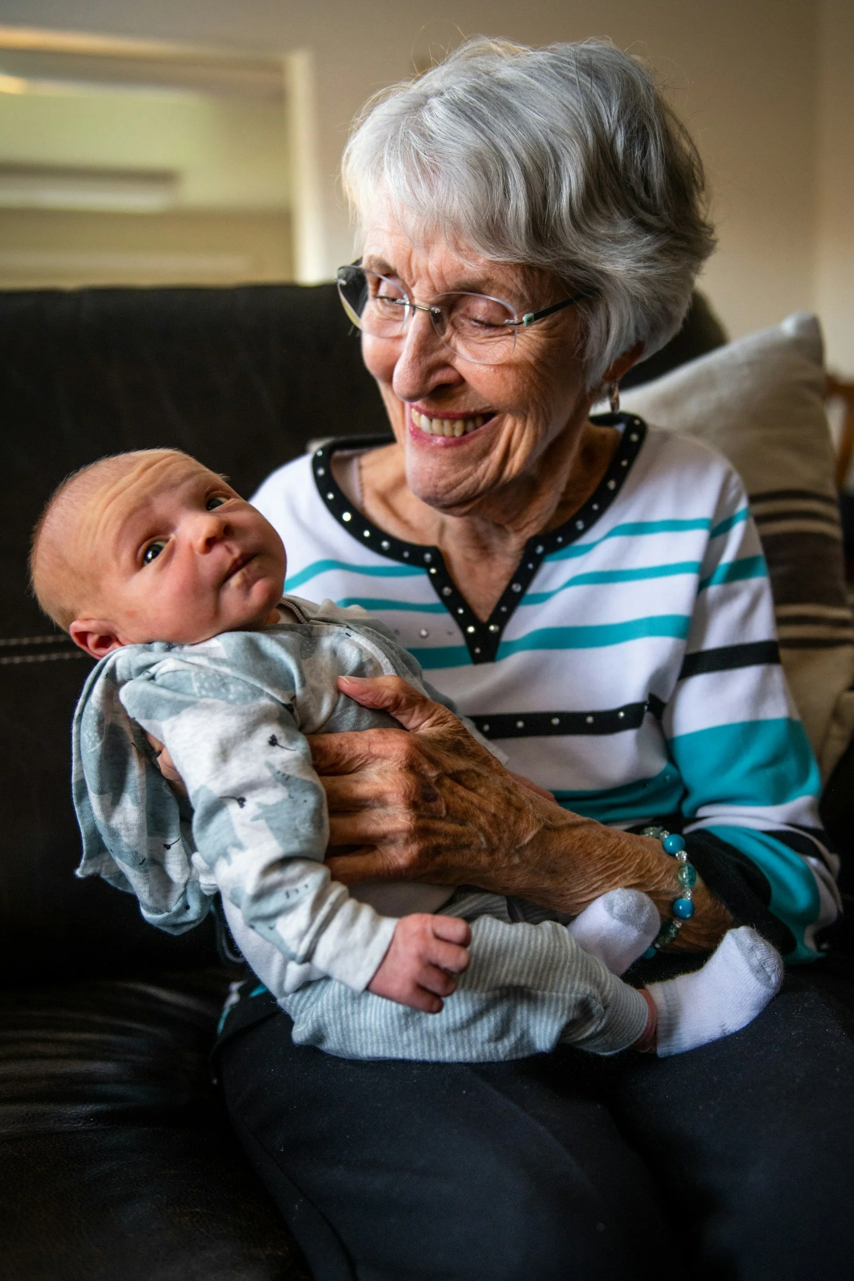 An elderly woman with short gray hair, glasses, and a striped shirt, smiling and holding a baby boy in her lap. The baby looks up at her with curiosity. They are sitting on a black leather sofa in a cozy room.