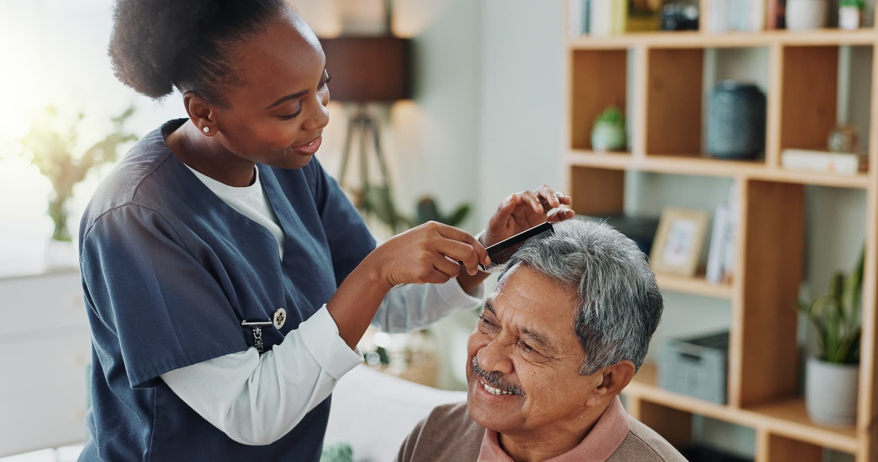 A nurse styles an elderly man's gray hair with a comb in a cozy, well-lit room with bookshelves and plants.