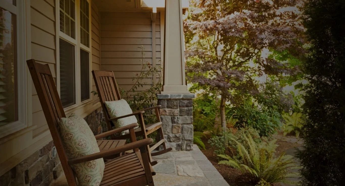 Two wooden rocking chairs with pillows on a porch next to a house, overlooking a garden with trees and ferns during sunset.