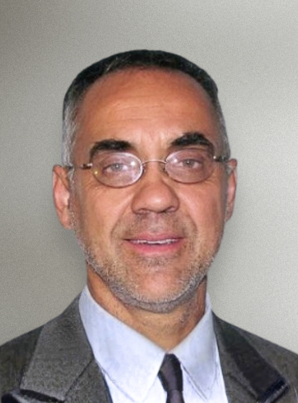 A middle-aged man with short dark hair, glasses, and a trimmed beard, dressed in a gray suit, white shirt, and black tie, standing against a plain background.