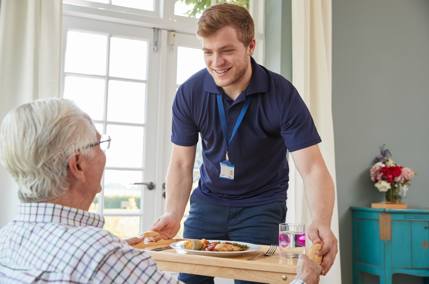 A young male caregiver with a blue shirt and an ID badge providing a meal to an elderly man with white hair and glasses, seated at a table with a plate of food, a glass of water, and utensils in a bright room with a door and flowers in the background.