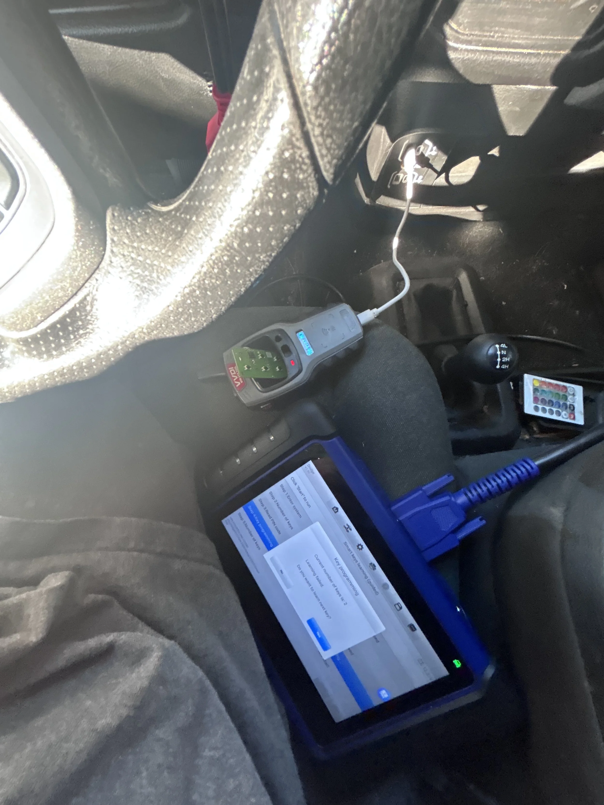 Inside a vehicle, a person is seated with a diagnostic scanner connected to the car's OBD port, a tablet displaying diagnostic information, and a light-emitting device with a green circuit board on the person's lap.