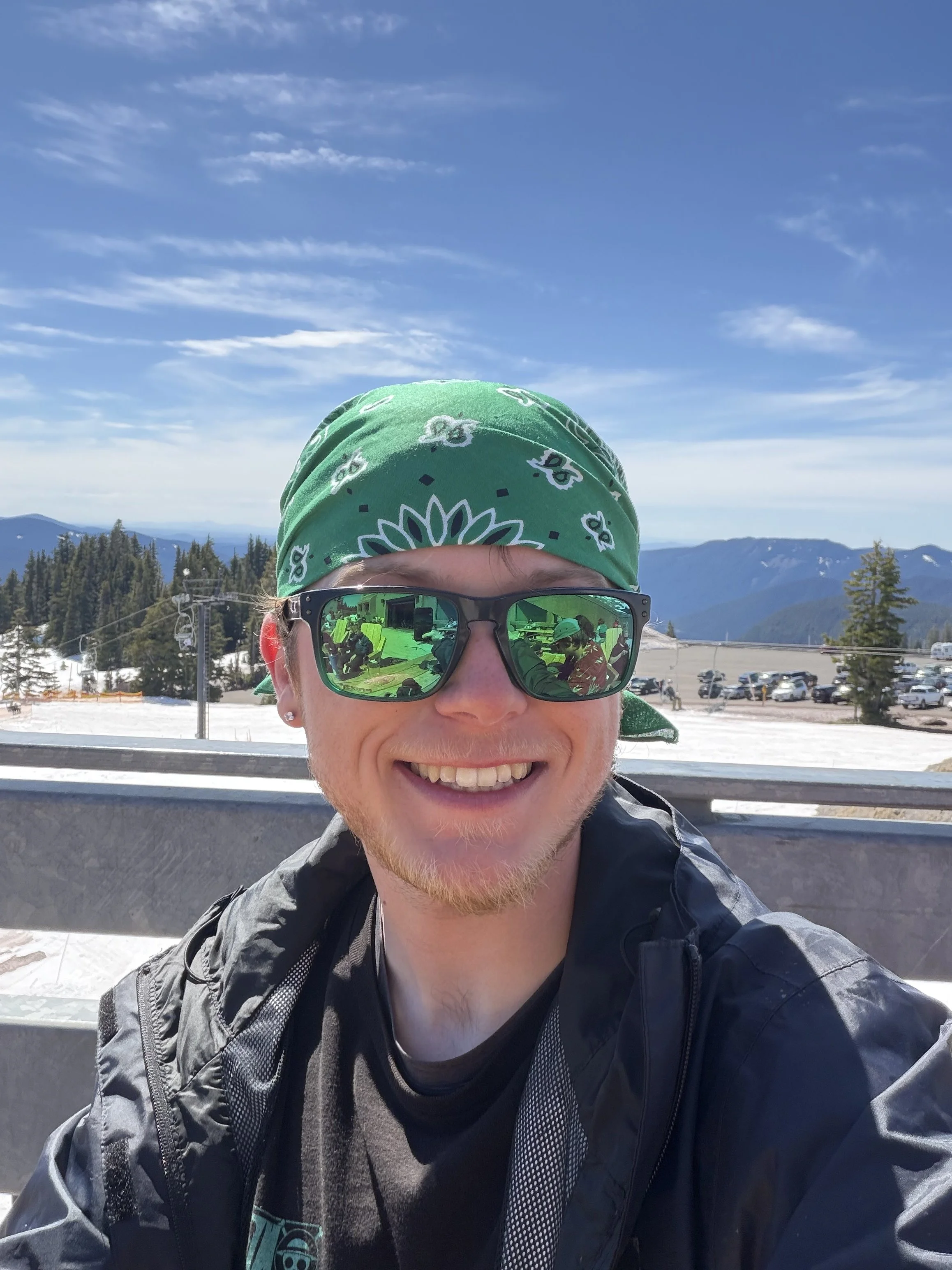 A person wearing sunglasses, a green bandana on their head, and a black jacket with a backpack, smiling at the camera outdoors with a mountain landscape and a parking lot in the background.