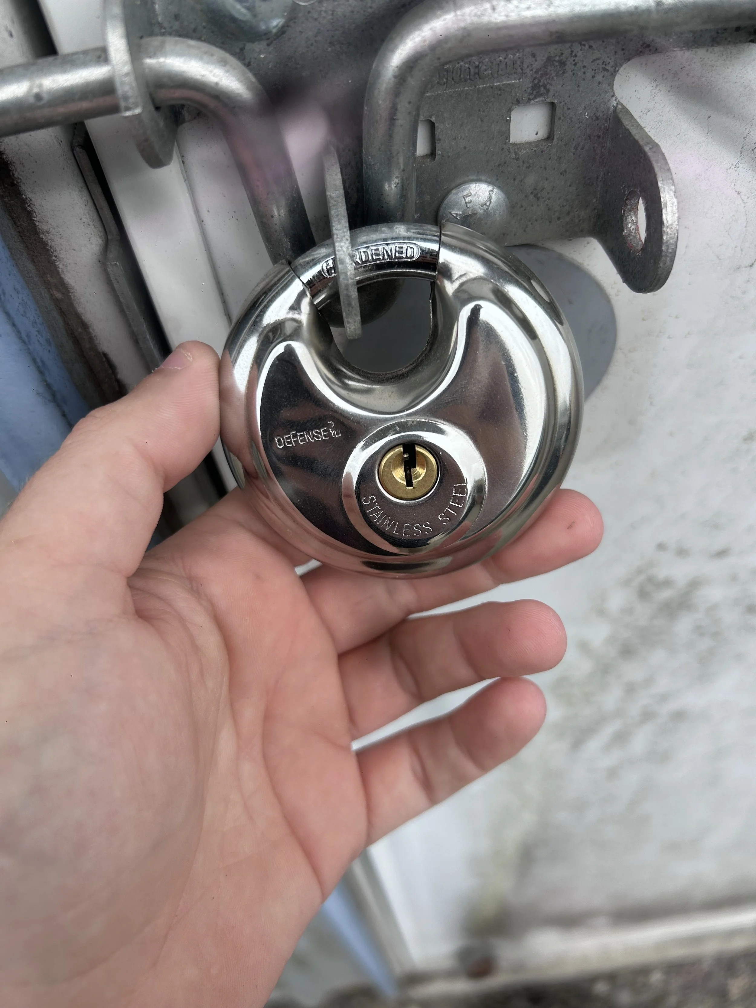 A person is holding a shiny, stainless steel padlock with the keyhole visible, securing it to a metal latch on a door or gate.